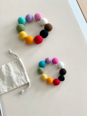 Two sets of colorful felt balls arranged in circles on white surface with small white drawstring bag nearby