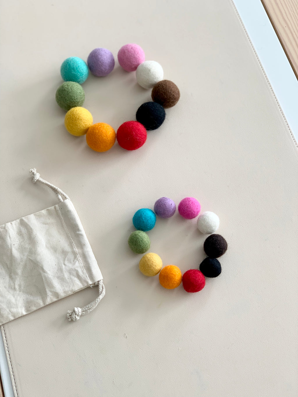 Two sets of colorful felt balls arranged in circles on white surface with small white drawstring bag nearby