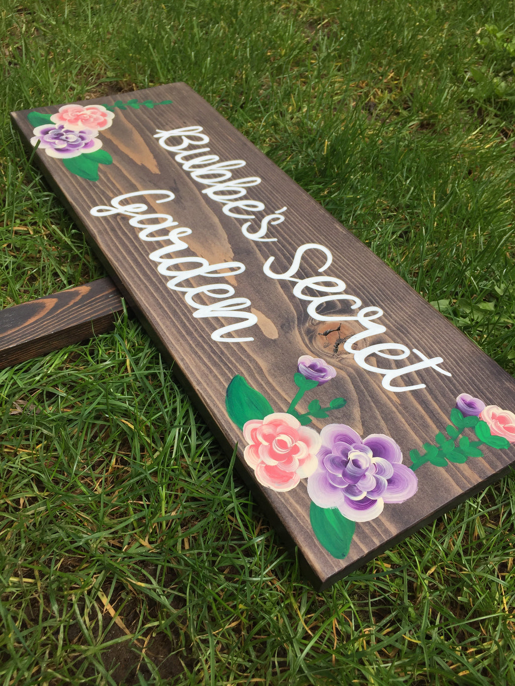 Large custom wooden garden sign with white cursive lettering and painted pink and purple floral accents on stained wood background