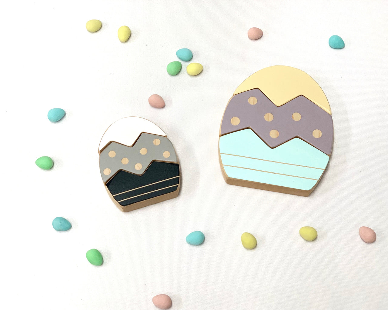 Two wooden Easter egg stacking toys with pastel colors and scattered small colorful candy eggs on white background