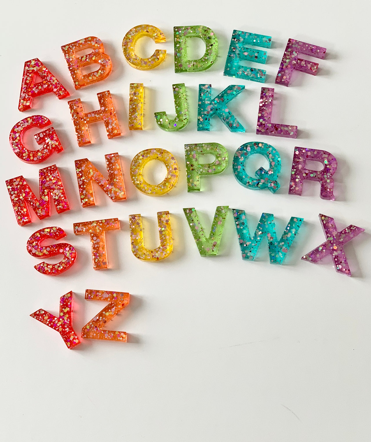Rainbow glitter resin uppercase alphabet letters arranged on white surface with sparkly details in multiple colors