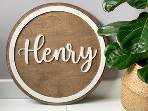 Round wooden name sign with white cursive letters and circular border on natural wood background next to green plant
