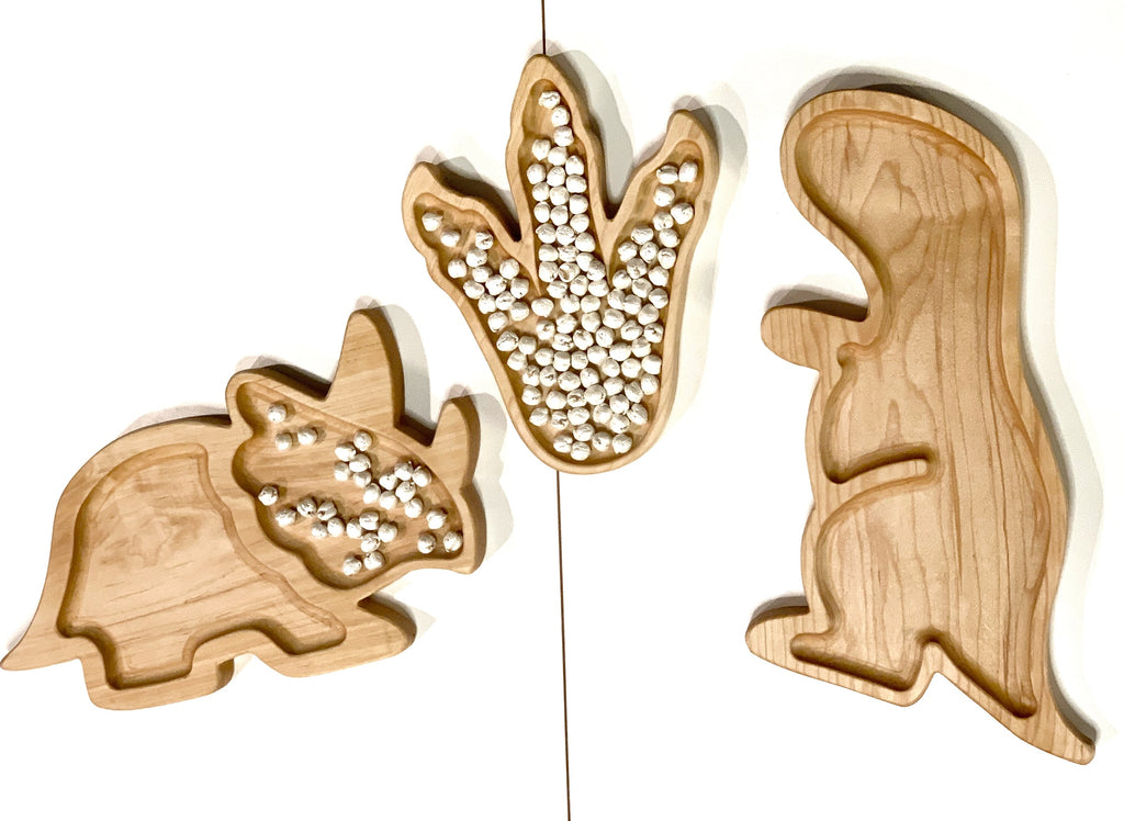 Set of three natural wooden sensory trays shaped like a triceratops, dinosaur footprint, and t-rex on white background