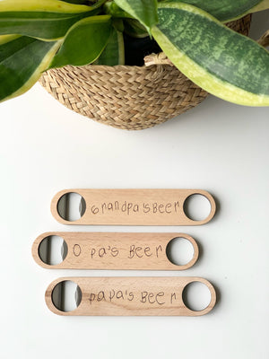Three custom wooden bottle openers with handwritten text laid flat on white surface below woven basket with green leaves