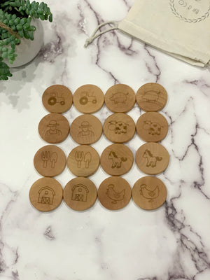Natural wooden round memory game pieces engraved with farm animals, tractor, farmer, barn, and utensils on marble surface near plant and pouch