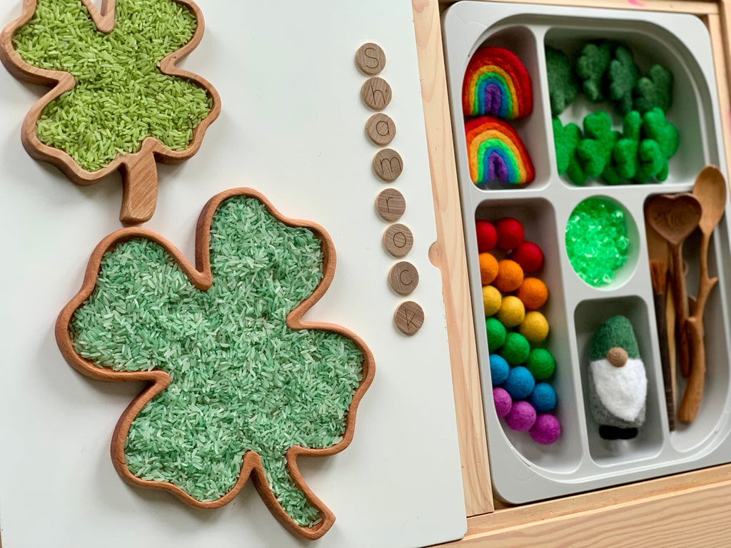 Small and large wooden shamrock sensory trays with green rice and rainbow felt balls in sectioned tray on wooden table