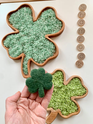 Wooden shamrock-shaped sensory trays filled with green rice and hand holding a small dark green felt clover toy