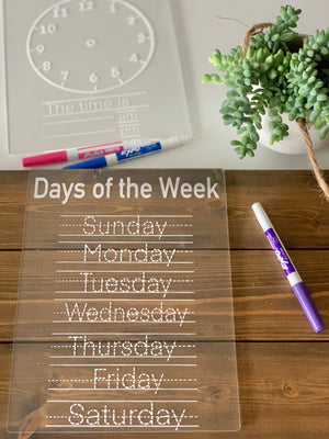 Clear acrylic tracing board with days of the week in white dashed letters on wood table with marker and plant nearby