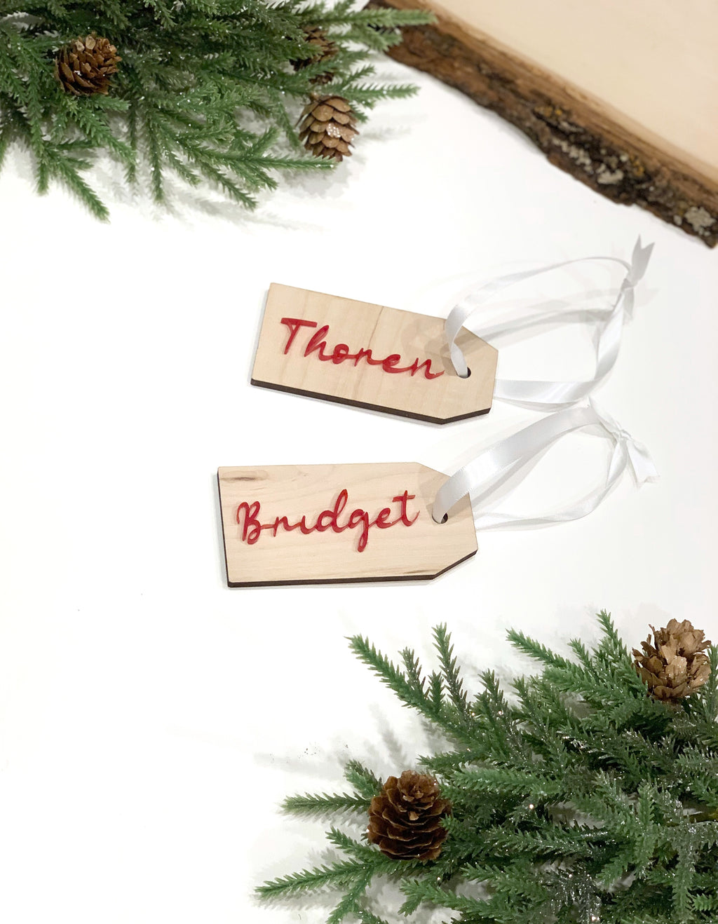 Custom wooden Christmas stocking and gift tags with red cursive names and white ribbon on white background with pine and pinecones