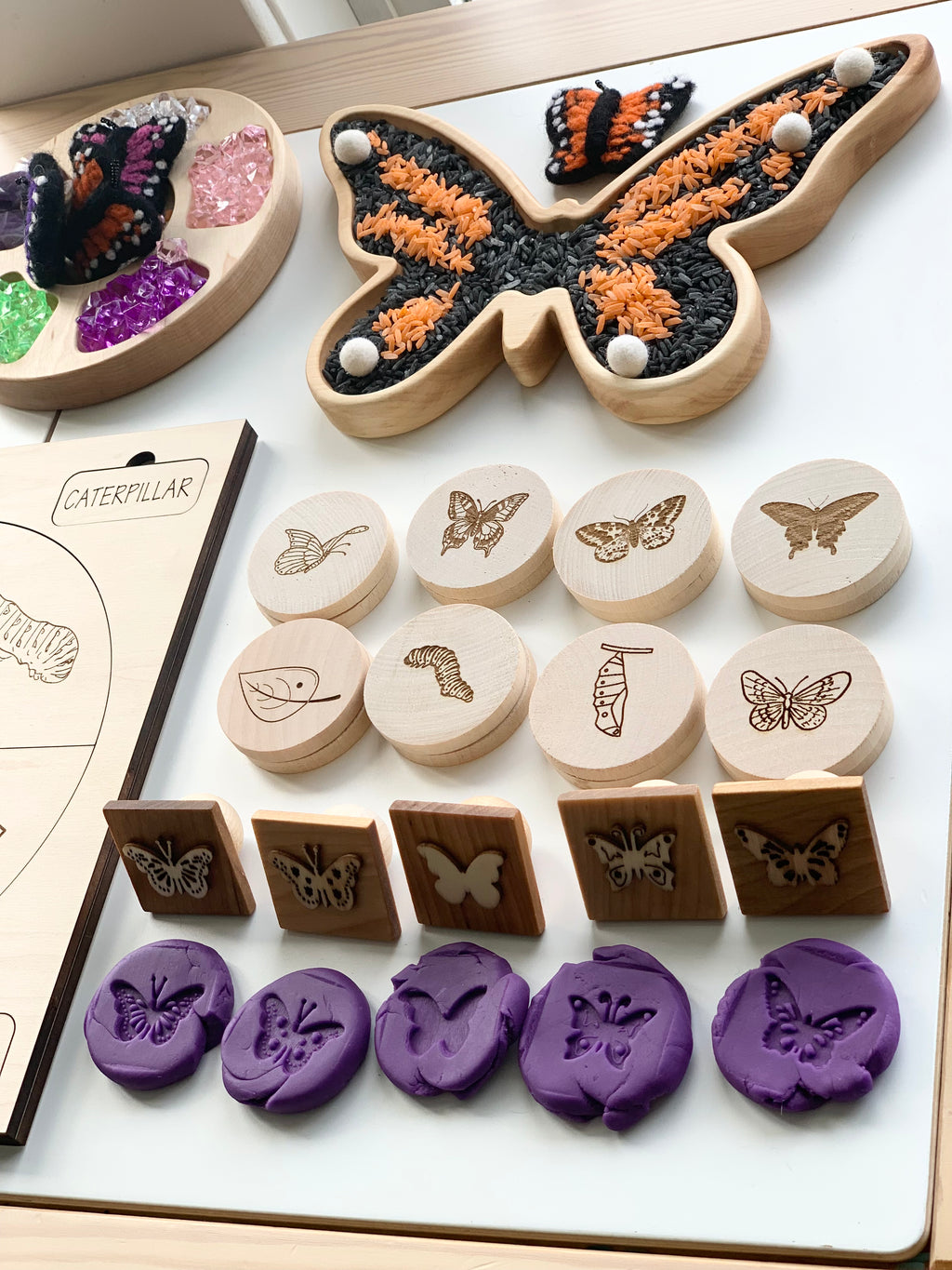 Wooden butterfly sensory tray filled with colored rice and set with wooden stamps and purple butterfly-shaped clay pieces on white surface