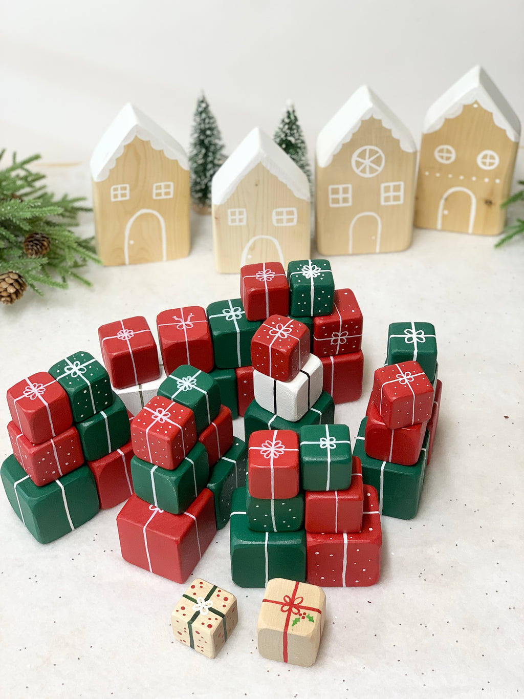 Stacked wooden Christmas gift blocks painted red, green, and white with white ribbon details and wooden snowy houses background
