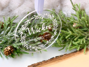 Clear acrylic Christmas ornament engraved with family name and decorative wreath surrounded by pine cones and greenery