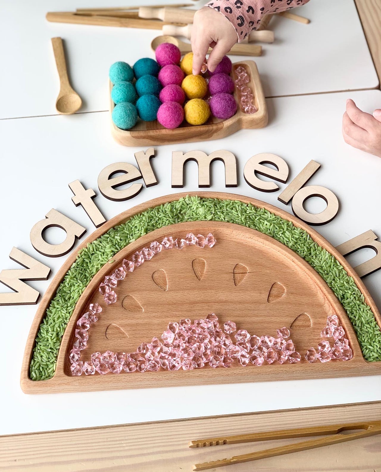 Large wooden watermelon sensory tray with engraved seeds, green rice border, and clear pink beads on light wood table