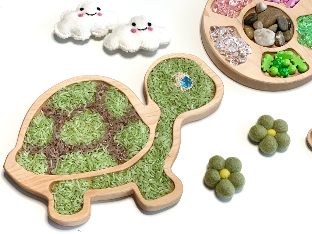 Turtle-shaped wooden sensory tray filled with green and brown rice and blue gem on white background