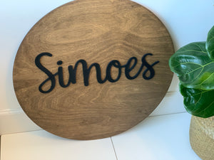 Round wooden 3D name sign with black cursive letters on natural wood background near green leafy plant on white floor