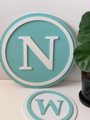 Round 3D monogram wooden sign with white letter N on turquoise background and decorative border next to small plant