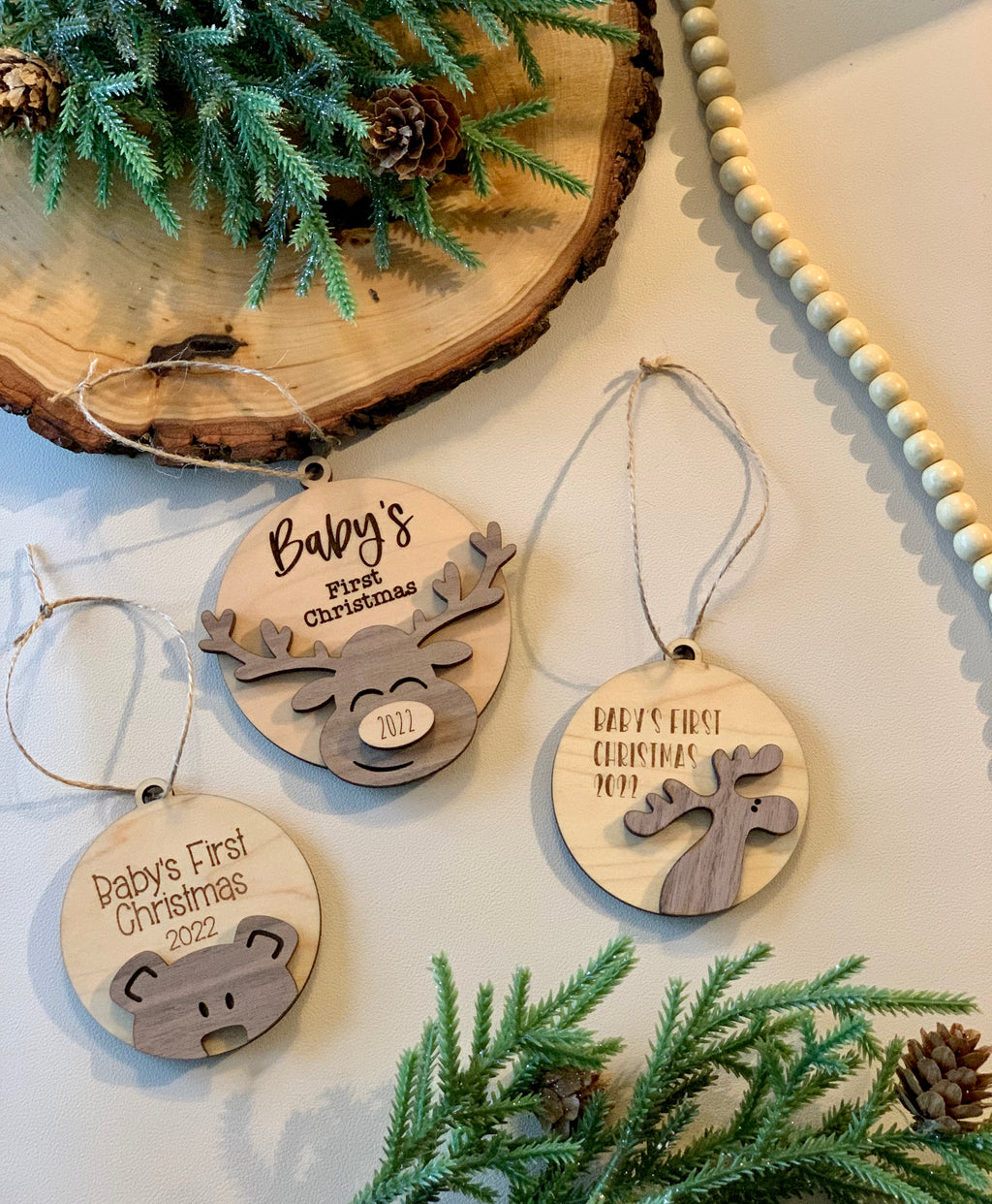 Three wooden baby’s first Christmas ornaments with moose and bear designs on white surface with greenery and pinecones