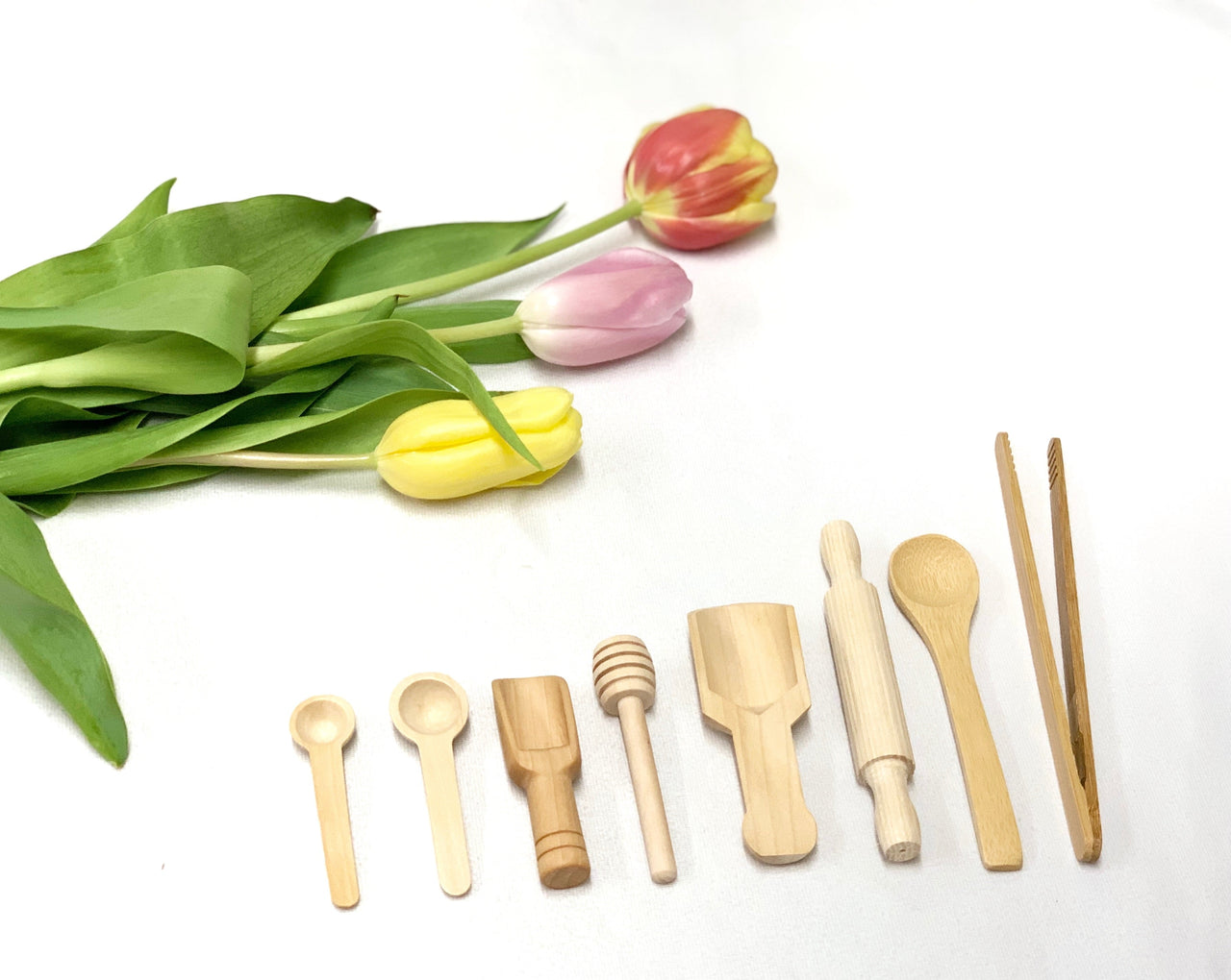 Small natural wooden sensory bin tools including spoons, scoop, honey dipper, rolling pin, spatula, and tongs on white surface with tulips
