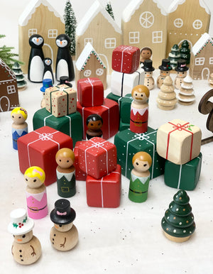 Hand-painted wooden peg dolls including Santa, elves, snowmen, penguins, surrounded by colorful gift boxes and Christmas trees on white surface