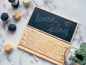 Wooden laptop toy with chalkboard screen displaying "learn + play" in chalk on marble surface with wooden blocks and plant