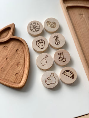 Wooden circular memory game discs with engraved simple fruit illustrations on white surface near carved wooden trays