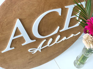 Large round wooden sign with white 3D block and cursive letters on natural wood background with flowers nearby