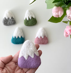 Handmade fair trade felt mountain in purple with white snowy peak held in hand with other colorful felt mountains nearby on white surface