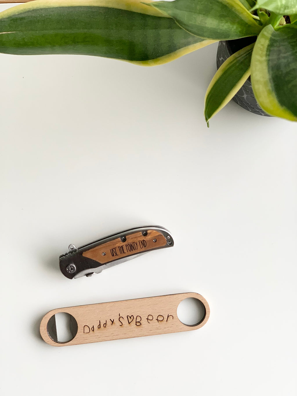 Custom wooden pocket knife with engraved handle next to wooden bottle opener with personalized engraving on white surface