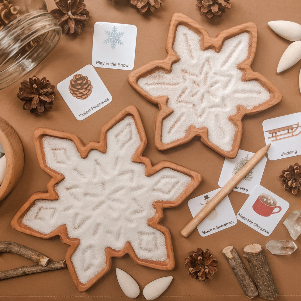 Natural wooden snowflake-shaped sensory trays filled with white sand and surrounded by winter-themed cards and pinecones on brown surface