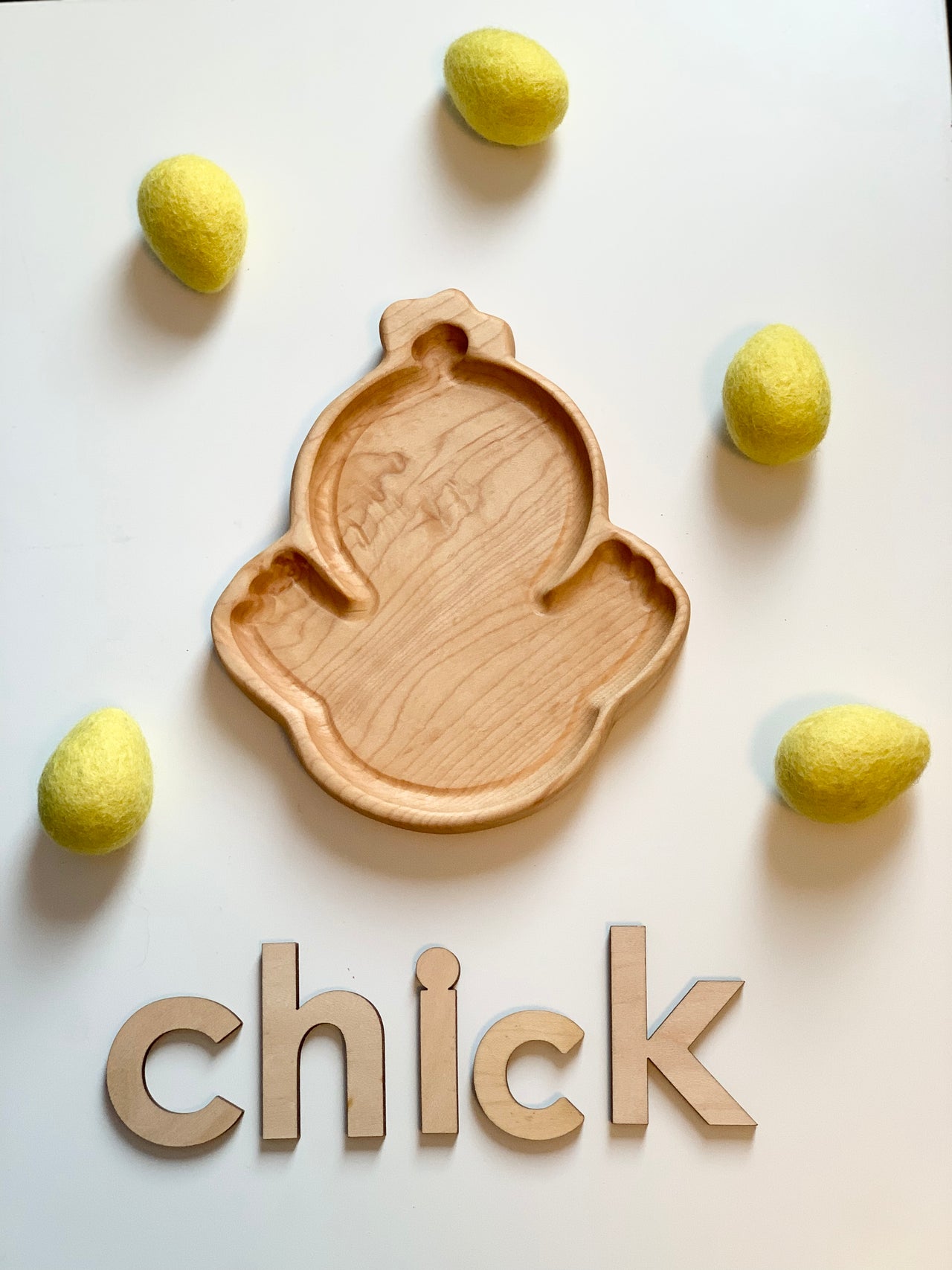 Wooden chick-shaped sensory tray surrounded by yellow felt eggs and wooden letters spelling chick on white surface