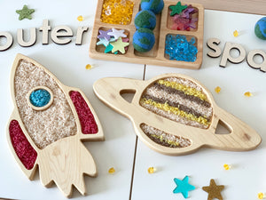Rectangle wooden tinker trays shaped like a rocket and planet filled with colored rice on white background with scattered stars