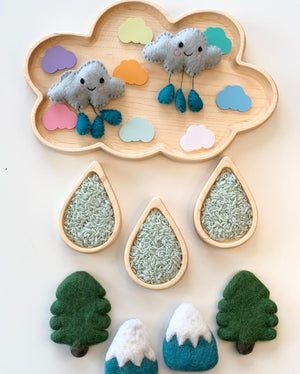 Natural wooden cloud-shaped sensory tray with colorful felt clouds, plush rain clouds, rice-filled raindrop bowls, and felt trees and mountains