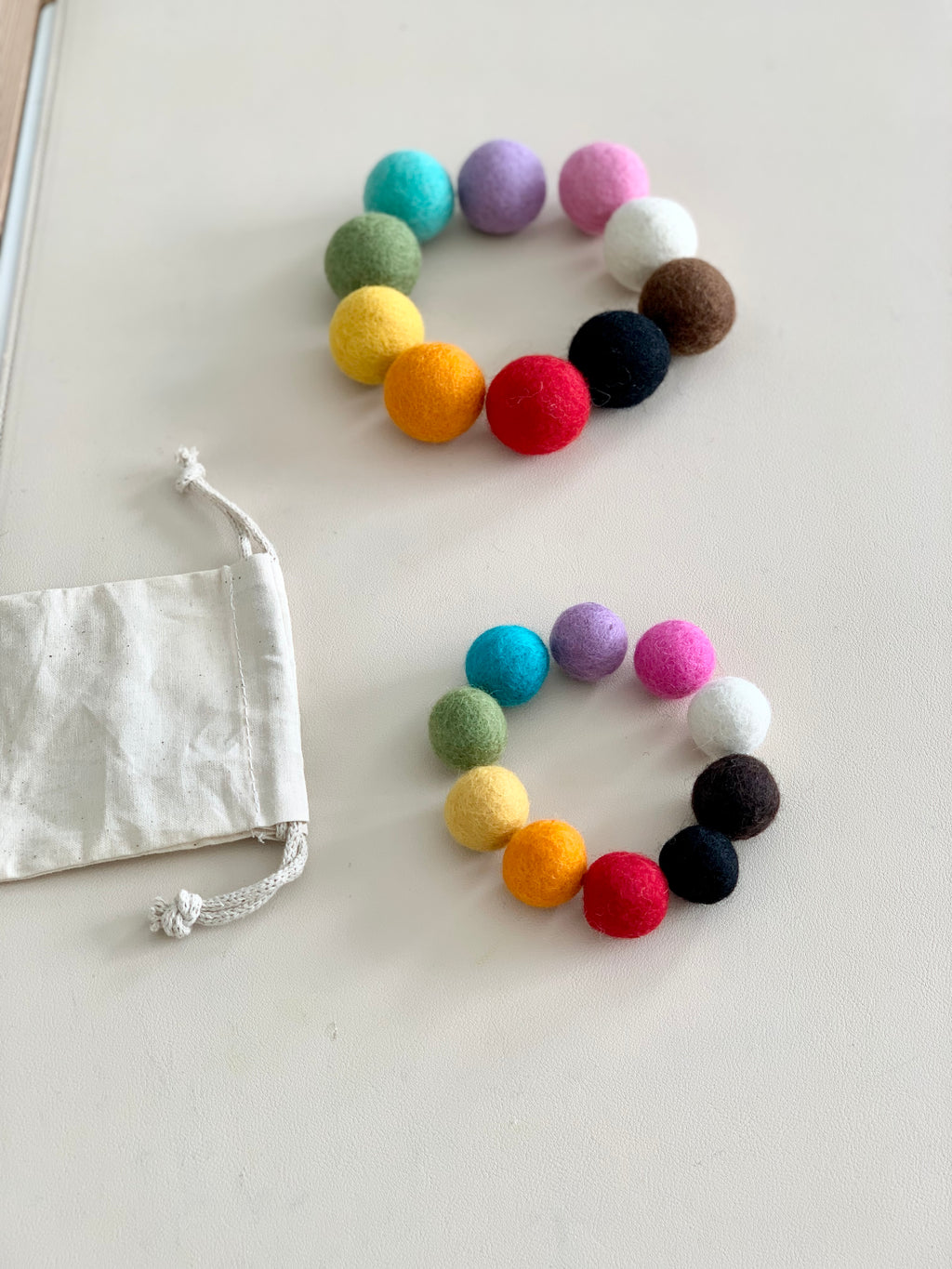 Two sets of colorful felt balls arranged in circles near a small white drawstring cotton bag on light surface