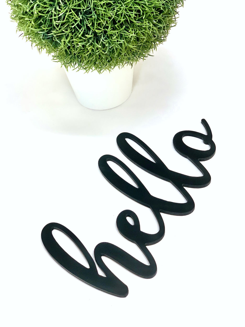 Black custom word cutout spelling hello in cursive script on white background next to small potted green plant