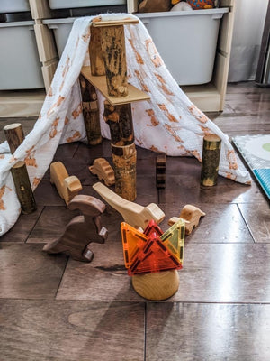 Assorted natural wood dinosaur toy figurines arranged in a play scene with wooden blocks and fabric tent on hardwood floor