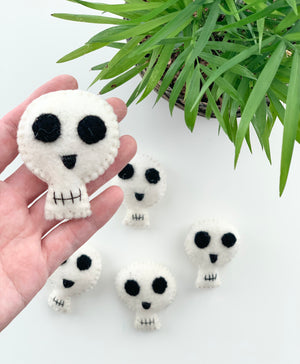 Handmade small white felt Halloween skull with black eyes and stitched mouth held by hand near green plant on white background
