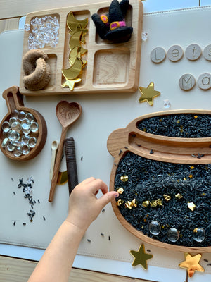 Small child’s hand playing with black dyed rice sensory bin filler with gold star confetti and wooden scoops on table