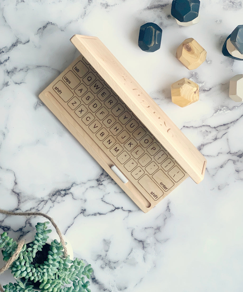 Wooden toy laptop with engraved keyboard and chalkboard screen on marble surface with wooden geometric shapes