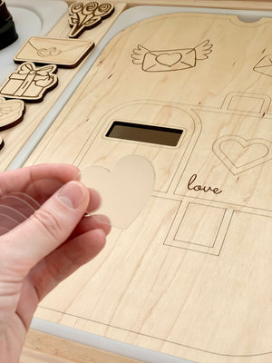 Hand holding translucent heart-shaped acrylic piece over wooden love-themed sensory play board with engraved shapes