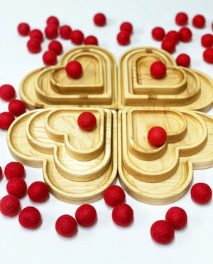 Natural wooden nesting heart plates arranged in clover shape surrounded by scattered small red felt balls on white background