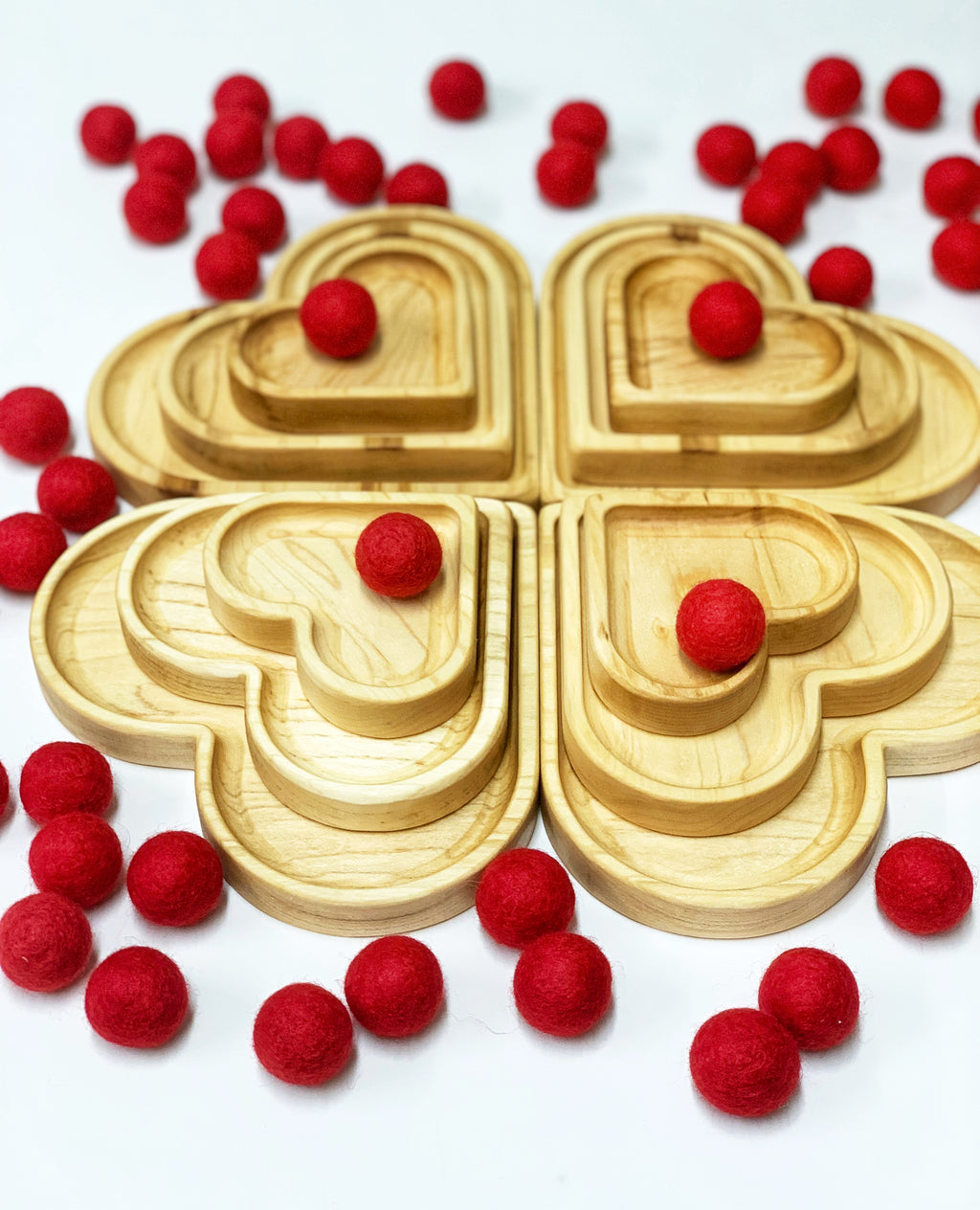 Natural wooden nesting heart plates arranged in clover shape surrounded by scattered small red felt balls on white background
