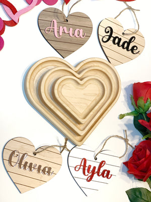 Four heart shaped wooden name tags with cursive letters in pink, black, silver, and red on a white background with red roses