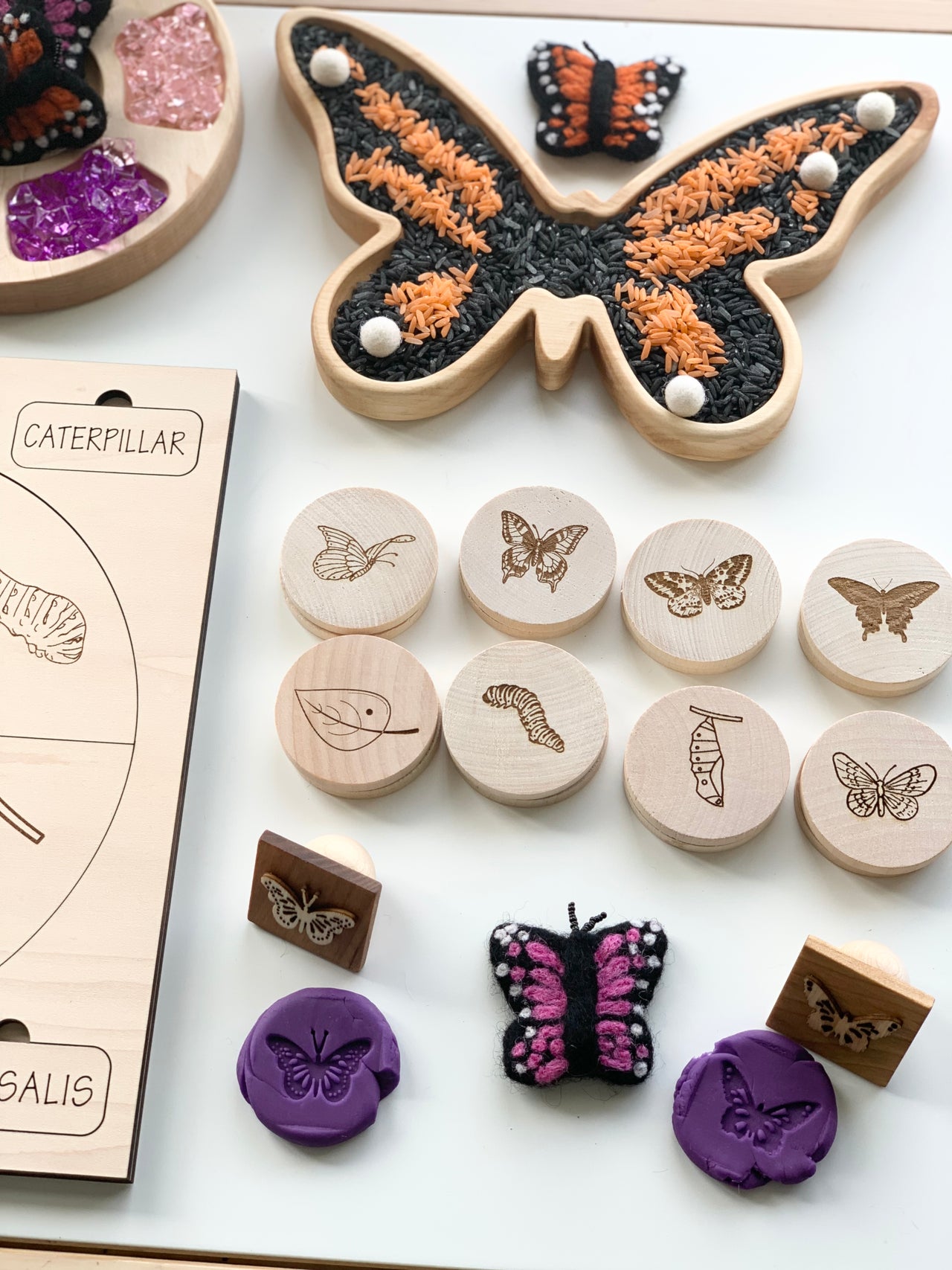 Wooden butterfly-shaped sensory tray filled with black and orange rice with wooden nature-themed stamps and butterfly crafts on white surface