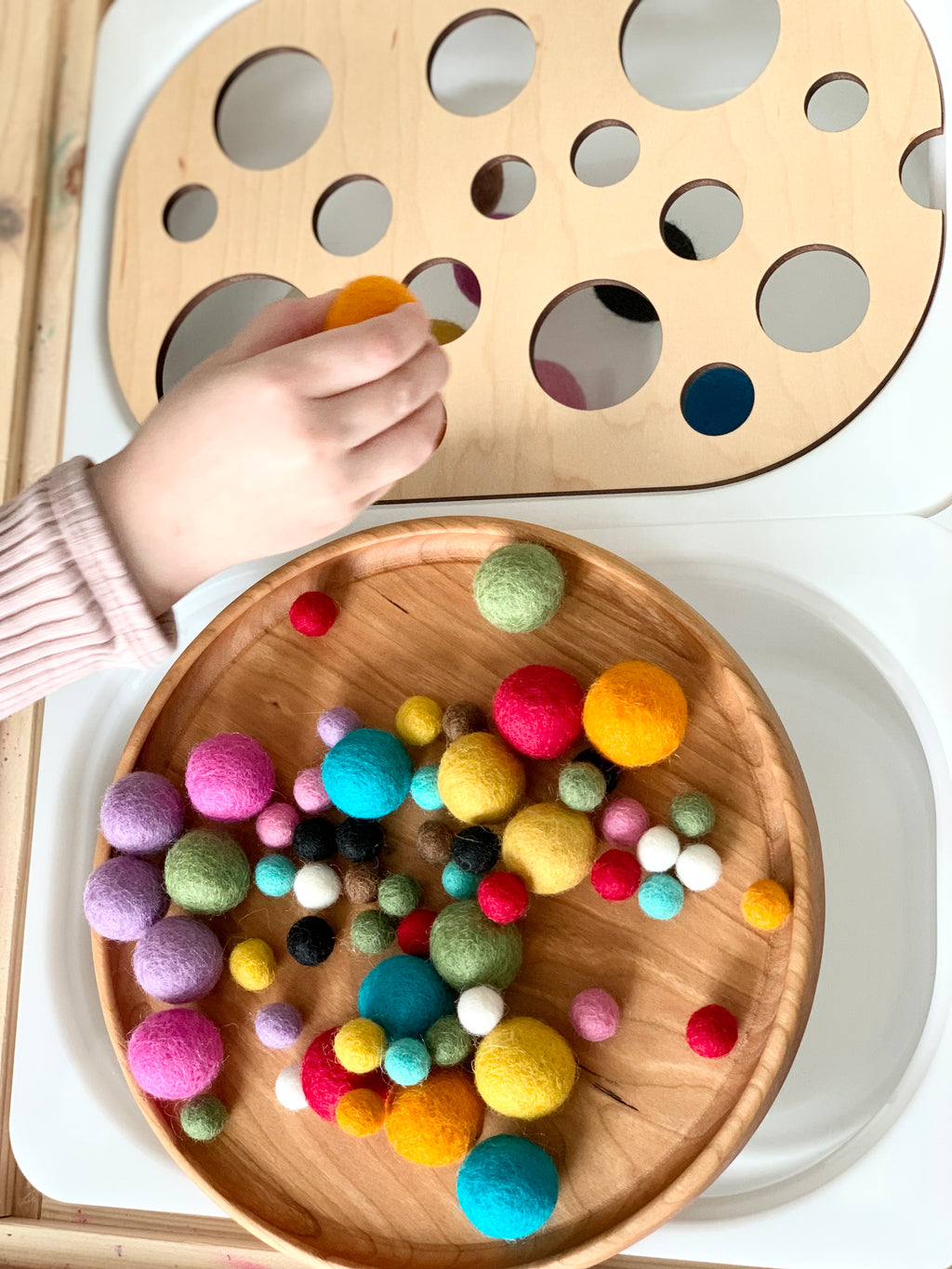 Child's hand placing a yellow felt ball into a wooden board with circular cutouts near a round tray of colorful felt balls