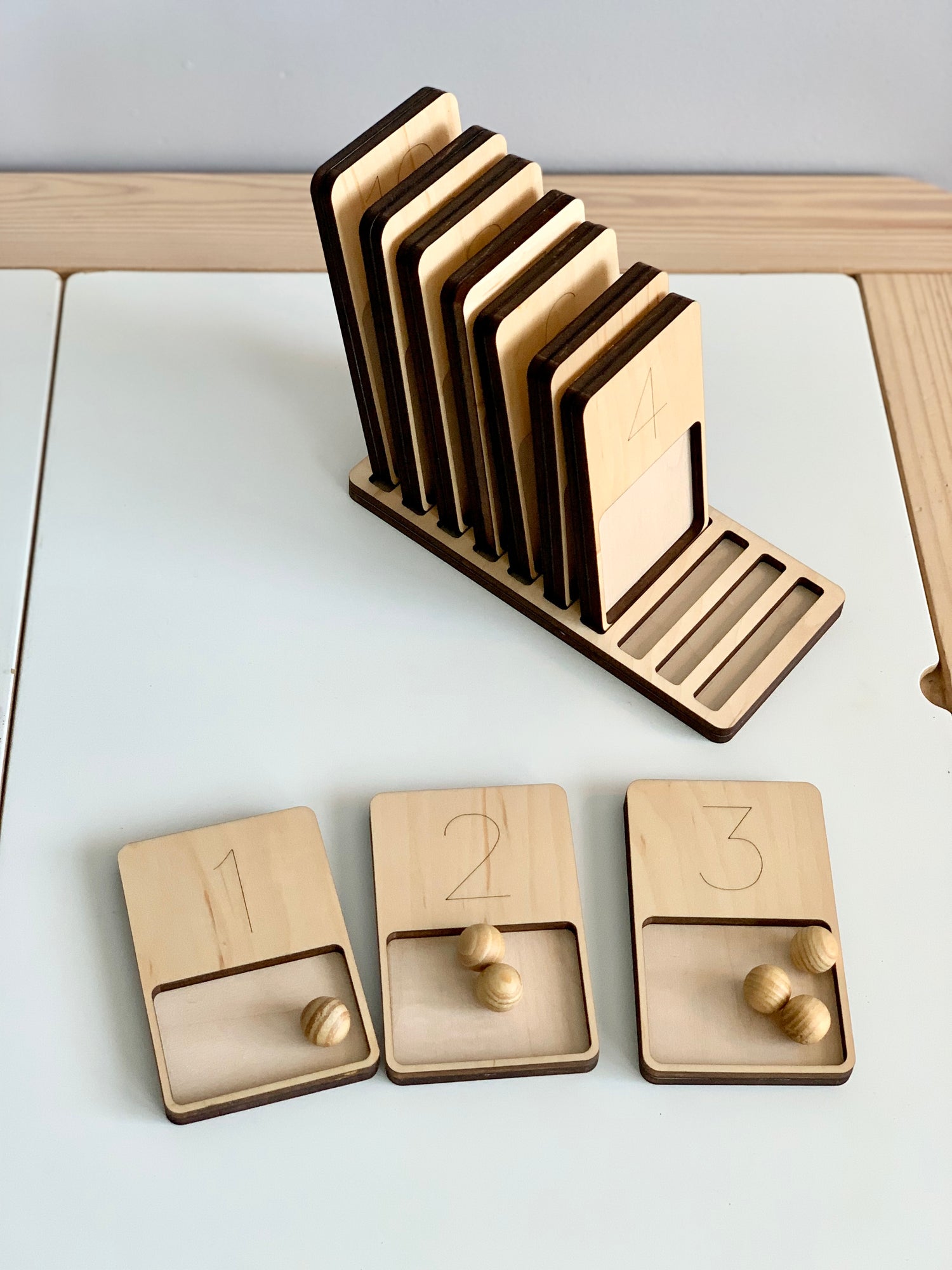 Wooden 1-10 counting trays with engraved numbers and wooden balls, arranged on a natural wood stand on white surface