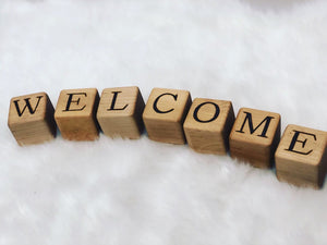 Natural wooden 1.5-inch blocks with engraved black letters spelling WELCOME arranged on white fabric background