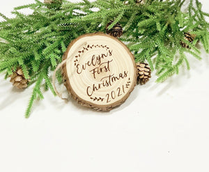 Wood burned wooden ornament engraved with Evelyn’s First Christmas 2021 surrounded by pine branches and pinecones on white background