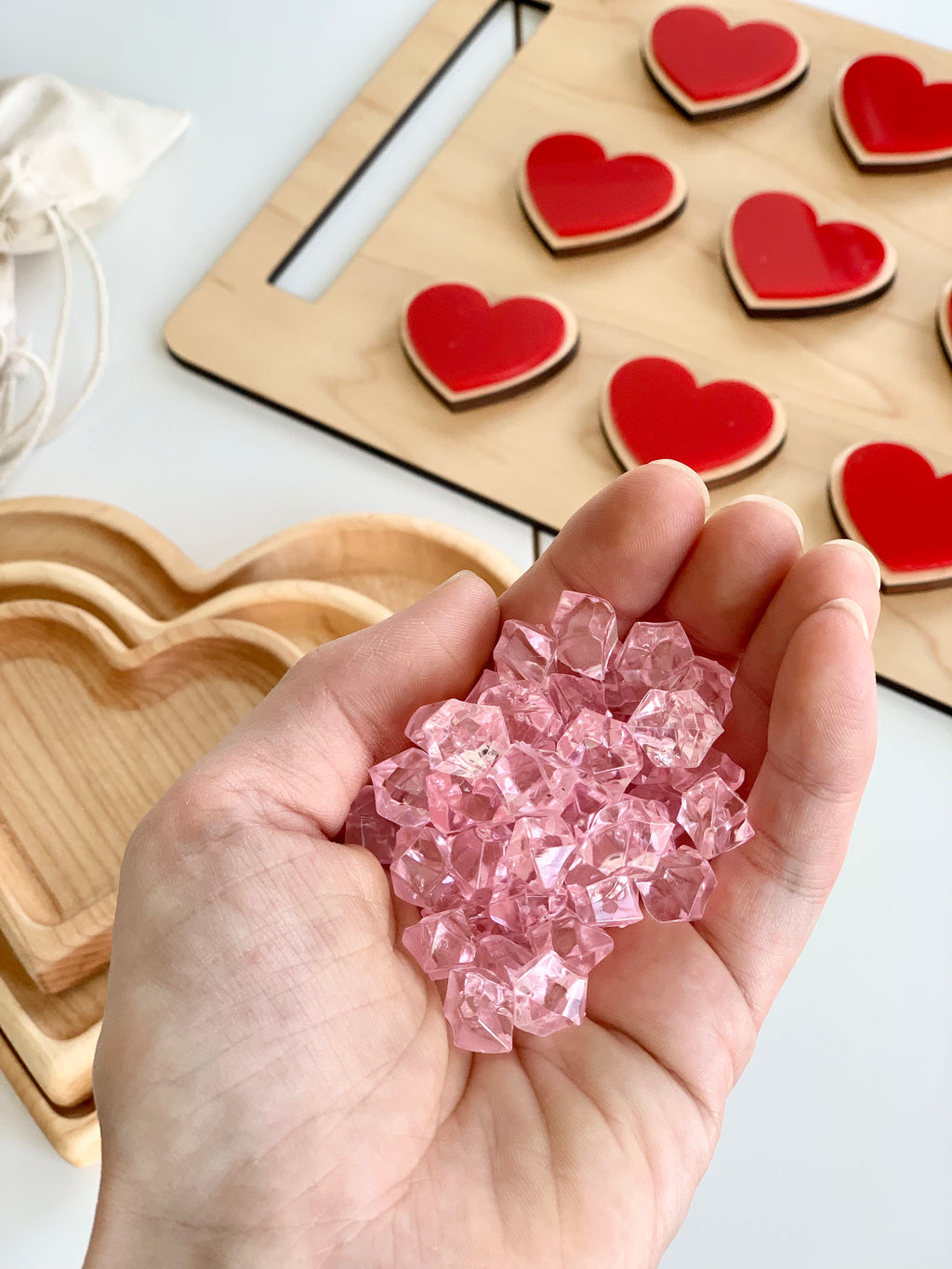 Hand holding multiple small pink acrylic treasure gems over wooden heart-shaped trays and a board with red wooden hearts
