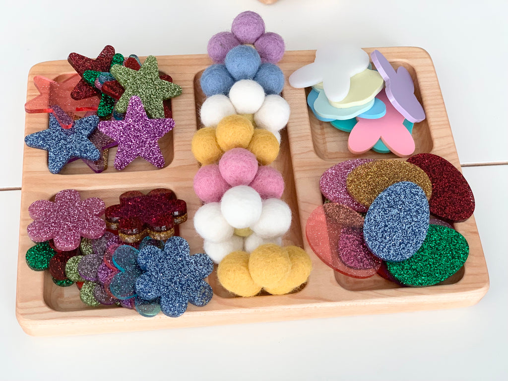 Natural wood rectangle tinker tray with colorful felt balls, glittery stars, flowers, and foam shapes arranged by compartment