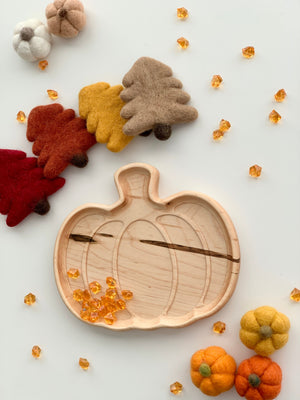 Light natural wooden pumpkin-shaped plate with engraved details surrounded by felt autumn leaves and pumpkins on white surface
